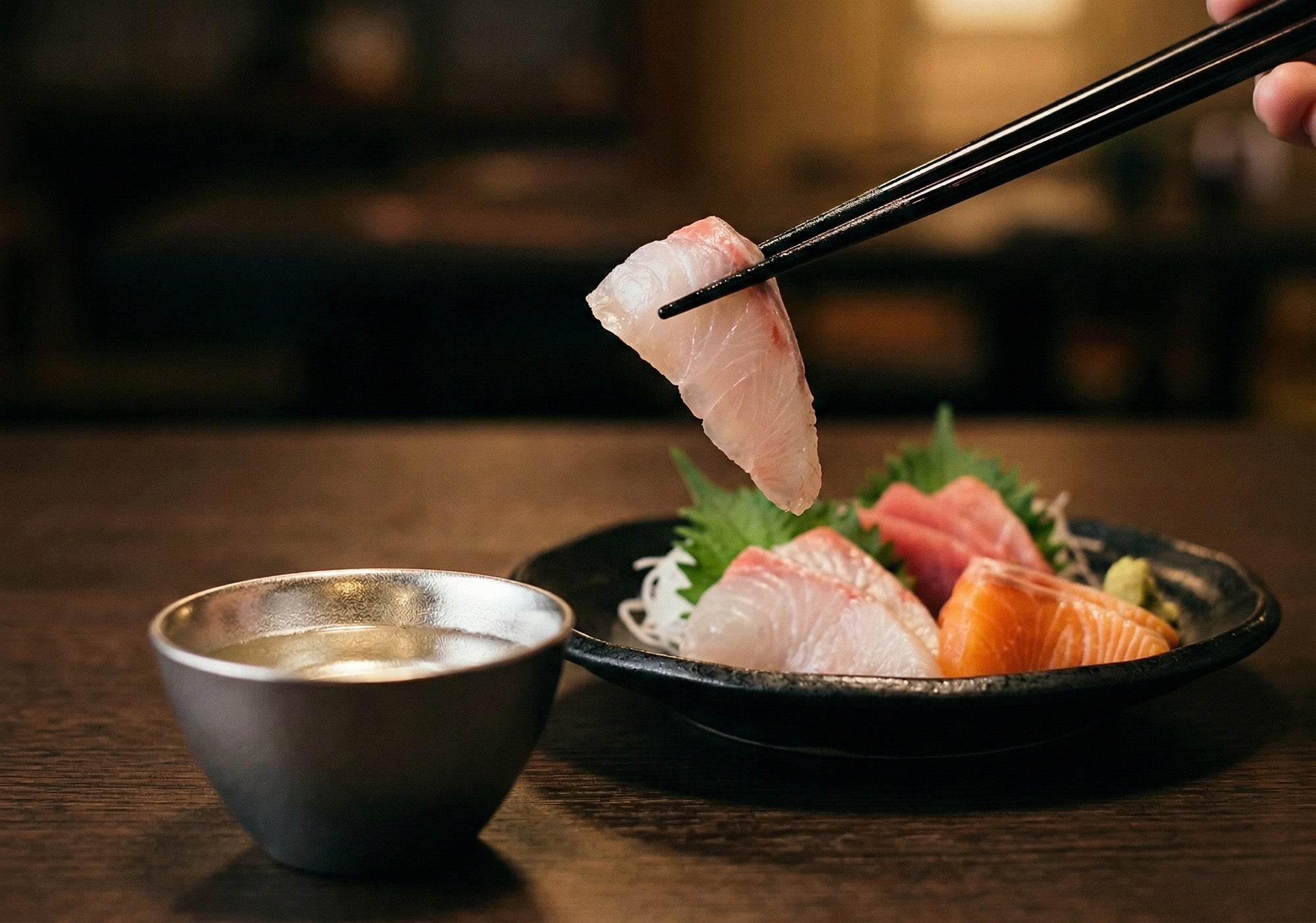 箸で白身の刺身をつまみ上げている瞬間。手前に酒が注がれた錫の盃