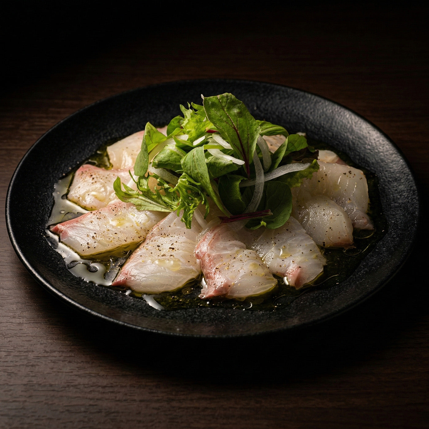 黒い皿に盛られた白身魚のカルパッチョ。オリーブオイルとベビーリーフ